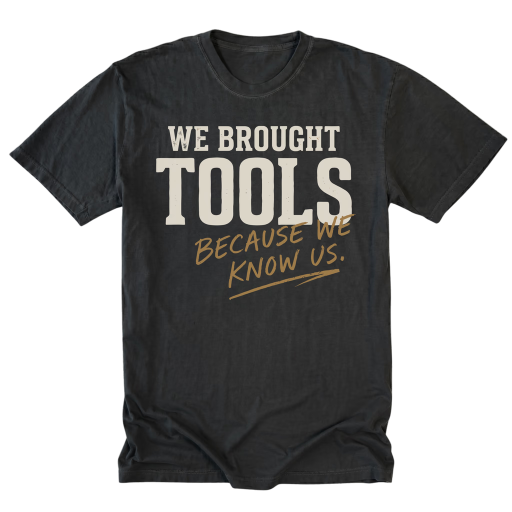 A black t-shirt with a graphic text print reading 'WE BROUGHT TOOLS BECAUSE WE KNOW US.' in cream and gold lettering, displayed flat against a white background.