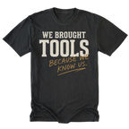 A black t-shirt with a graphic text print reading 'WE BROUGHT TOOLS BECAUSE WE KNOW US.' in cream and gold lettering, displayed flat against a white background.