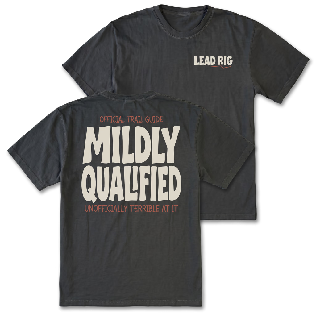 Two black graphic t-shirts shown on a white background, with the front and back views overlapping. The front features a small 'LEAD RIG' logo on the left chest, and the back features a large graphic print reading 'OFFICIAL TRAIL GUIDE MILDLY QUALIFIED UNOFFICIALLY TERRIBLE AT IT' in cream and orange lettering.