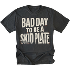 A black graphic t-shirt with the text 'BAD DAY TO BE A SKID PLATE' printed in distressed off-white lettering with small orange road-striping accents, shown on a plain white background.