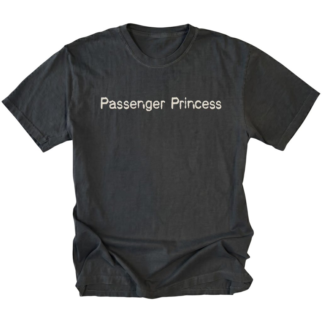 A black t-shirt with the words 'Passenger Princess' printed in a light beige serif font across the chest, displayed flat against a solid white background.