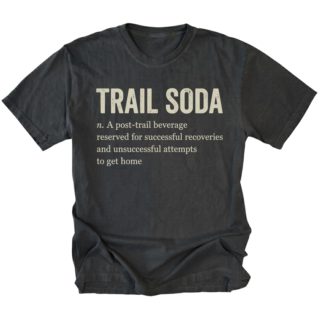 A black t-shirt with a humorous definition of 'Trail Soda' printed in beige text on the front, displayed against a solid white background.