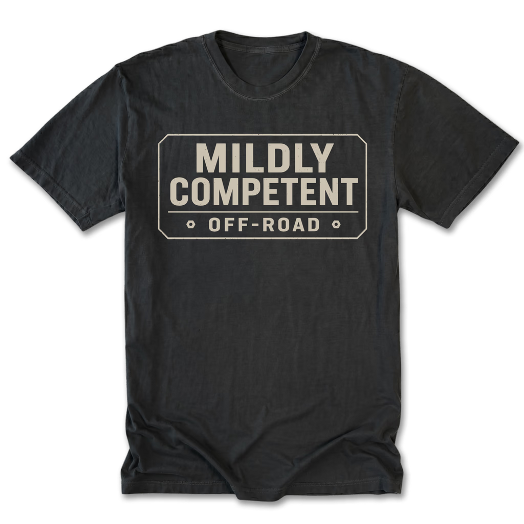A black tall tee crewneck t-shirt with a distressed beige graphic on the chest that reads 'MILDLY COMPETENT OFF-ROAD' inside a road-sign style border, shown on a plain white background.