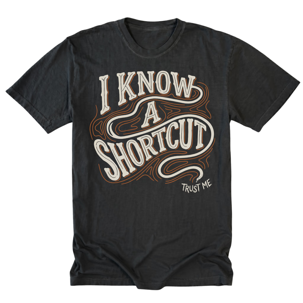 A black graphic t-shirt featuring the text 'I KNOW A SHORTCUT' in a stylized cream and brown outline font, with 'TRUST ME' printed below it.