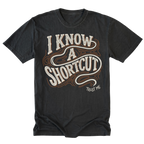 A black graphic t-shirt featuring the text 'I KNOW A SHORTCUT' in a stylized cream and brown outline font, with 'TRUST ME' printed below it.