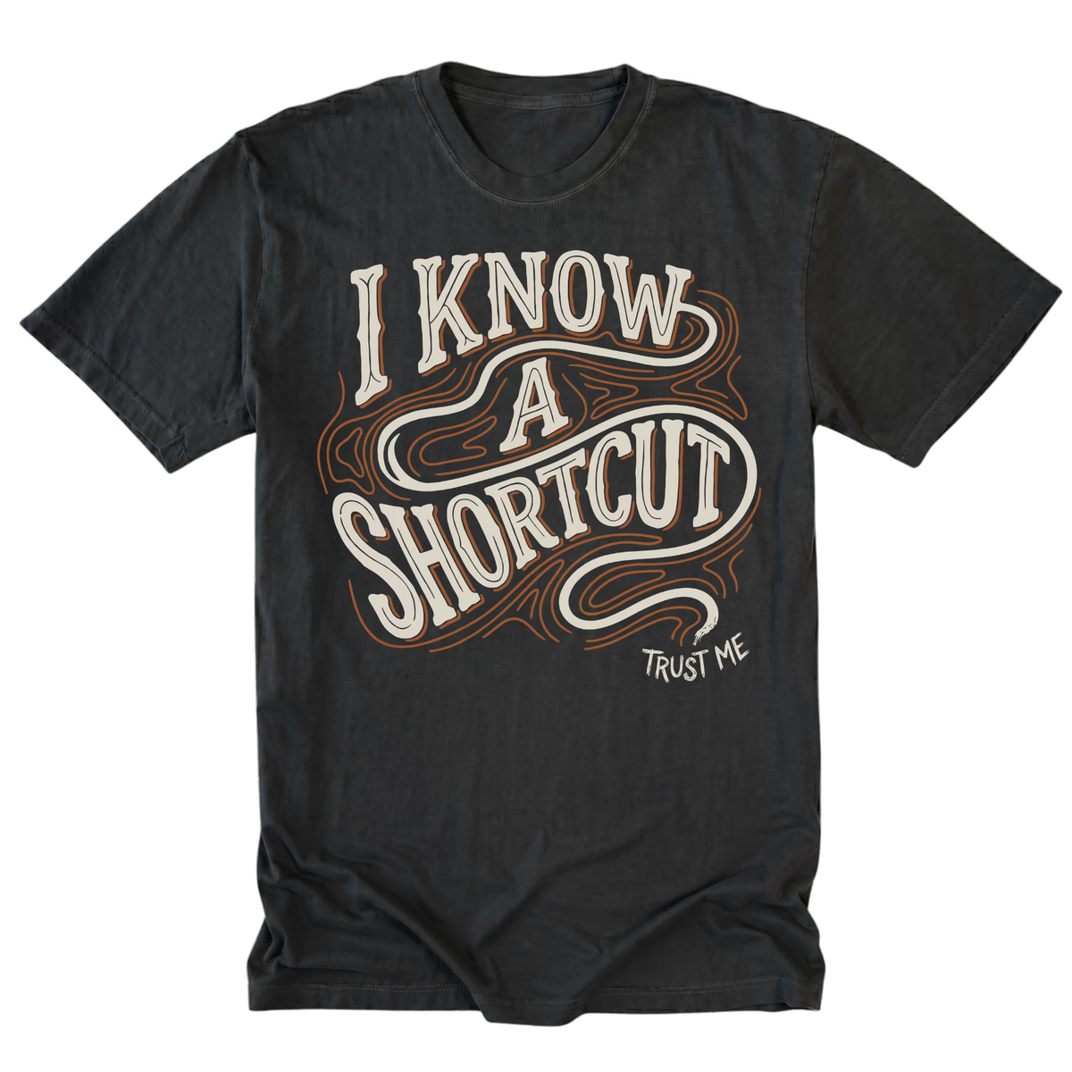 A black graphic t-shirt featuring the text 'I KNOW A SHORTCUT' in a stylized cream and brown outline font, with 'TRUST ME' printed below it.