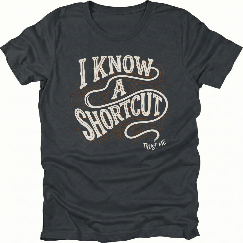Black t-shirt with vintage typography reading I Know A Shortcut Trust Me with winding trail lines, front view by Trail Snark.