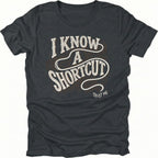 Black t-shirt with vintage typography reading I Know A Shortcut Trust Me with winding trail lines, front view by Trail Snark.