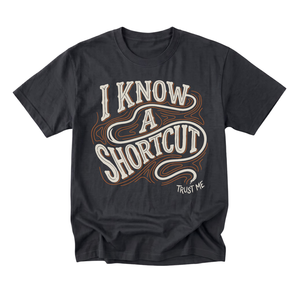 A black graphic t-shirt featuring the phrase 'I KNOW A SHORTCUT' in a retro, wavy cream and brown font, with 'TRUST ME' printed in small white text below it, displayed on a clean white background.