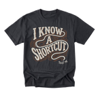 A black graphic t-shirt featuring the phrase 'I KNOW A SHORTCUT' in a retro, wavy cream and brown font, with 'TRUST ME' printed in small white text below it, displayed on a clean white background.