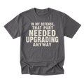 A charcoal gray crewneck t-shirt with a distressed cream-colored text graphic on the front that reads 'IN MY DEFENSE, THAT PART NEEDED UPGRADING ANYWAY'. The t-shirt is displayed flat against a solid white background.