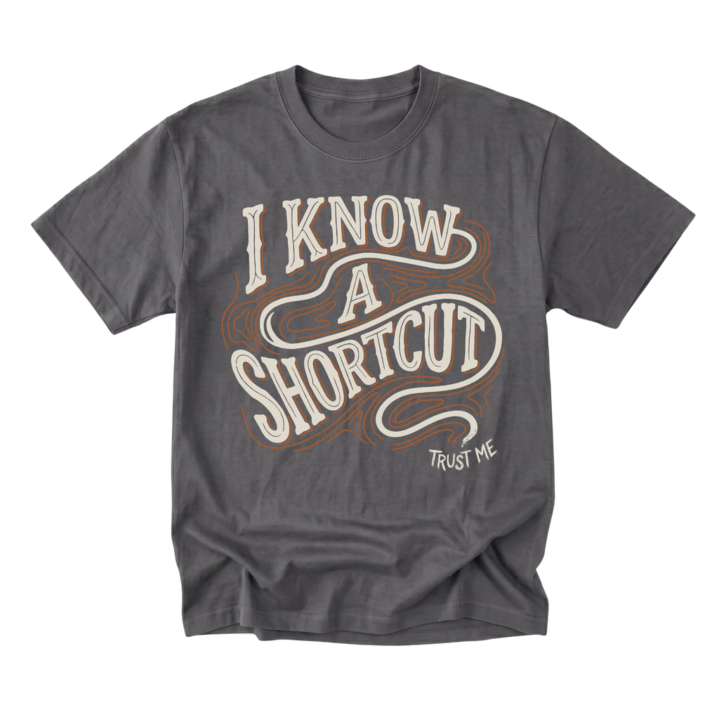 A charcoal gray graphic t-shirt featuring the phrase 'I KNOW A SHORTCUT' in a retro cream and brown wavy font, with 'TRUST ME' printed in small white text below, displayed on a clean white background.