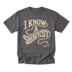 A charcoal gray graphic t-shirt featuring the phrase 'I KNOW A SHORTCUT' in a retro cream and brown wavy font, with 'TRUST ME' printed in small white text below, displayed on a clean white background.