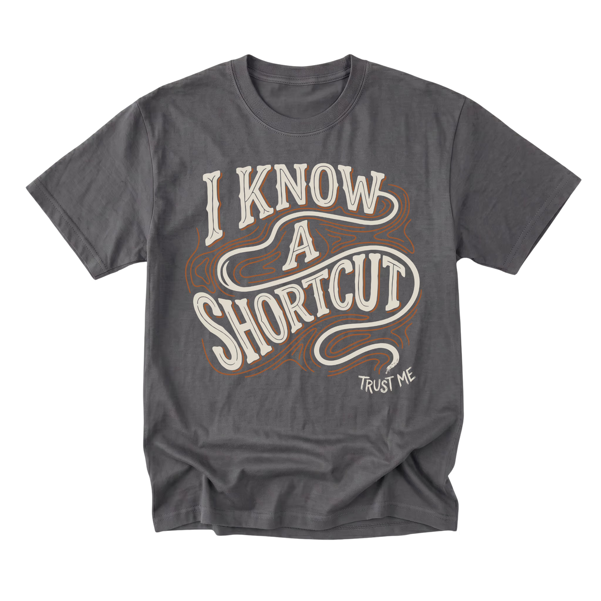 A charcoal gray graphic t-shirt featuring the phrase 'I KNOW A SHORTCUT' in a retro cream and brown wavy font, with 'TRUST ME' printed in small white text below, displayed on a clean white background.