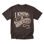 A dark brown short-sleeve t-shirt with a retro-style graphic reading 'I KNOW A SHORTCUT' in white and orange wavy lettering, with 'TRUST ME' printed below on the right side.