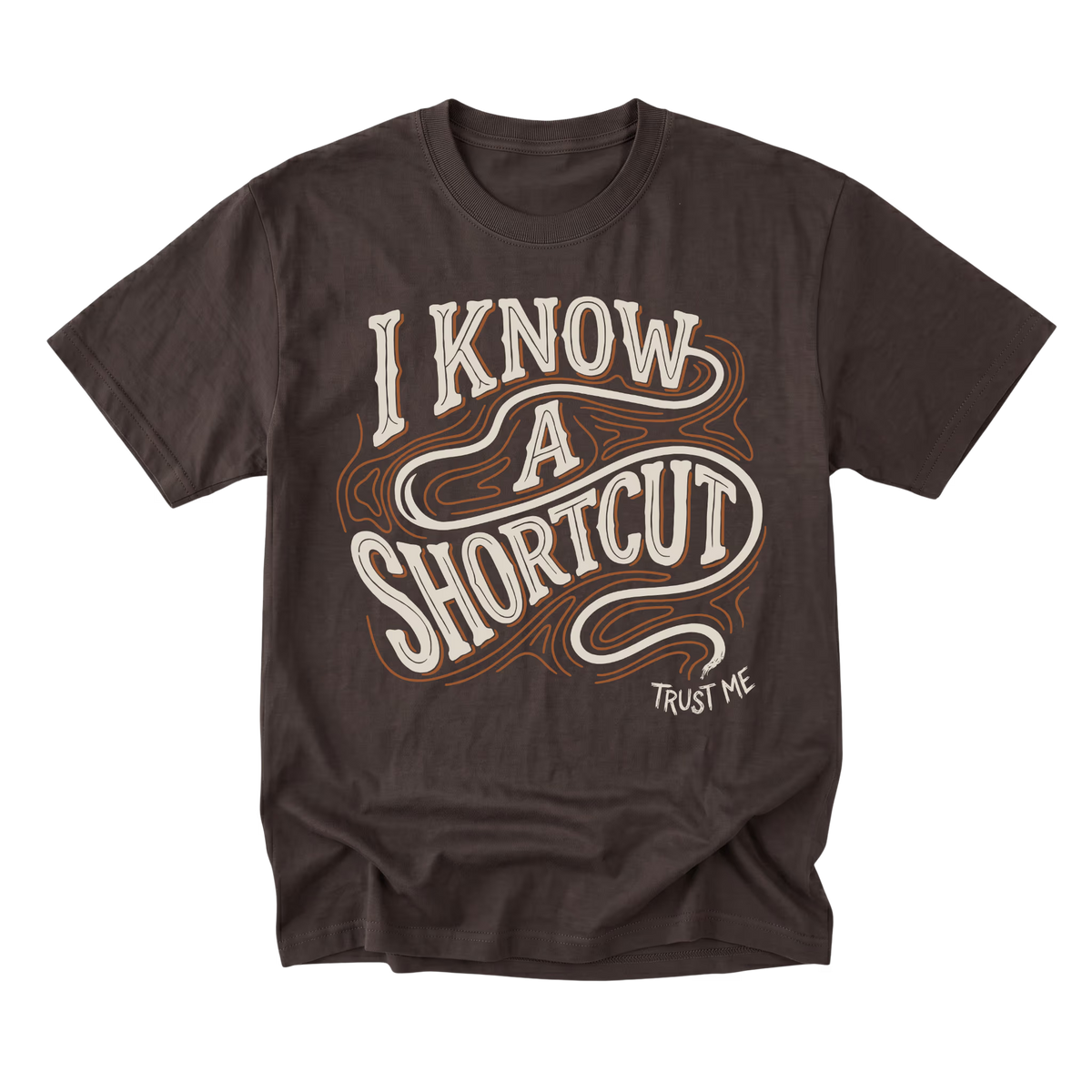 A dark brown short-sleeve t-shirt with a retro-style graphic reading 'I KNOW A SHORTCUT' in white and orange wavy lettering, with 'TRUST ME' printed below on the right side.