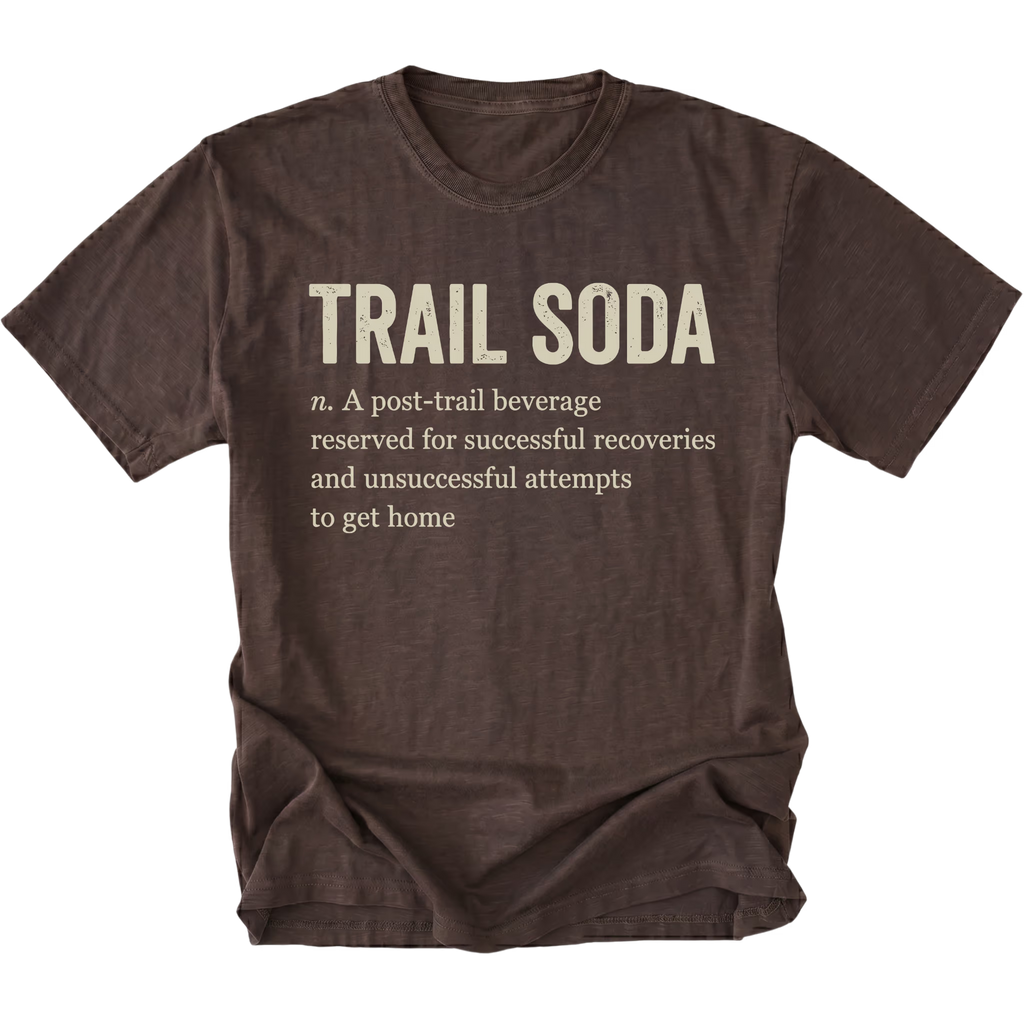 A dark brown short-sleeve t-shirt with a distressed texture, featuring a humorous definition of 'Trail Soda' printed in beige text on the front.