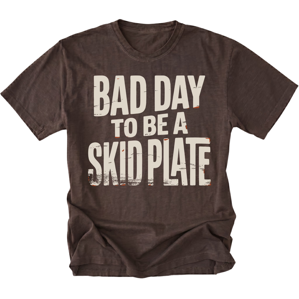 A dark brown short-sleeve t-shirt with a distressed white graphic text reading 'BAD DAY TO BE A SKID PLATE' on the front, displayed against a solid white background.