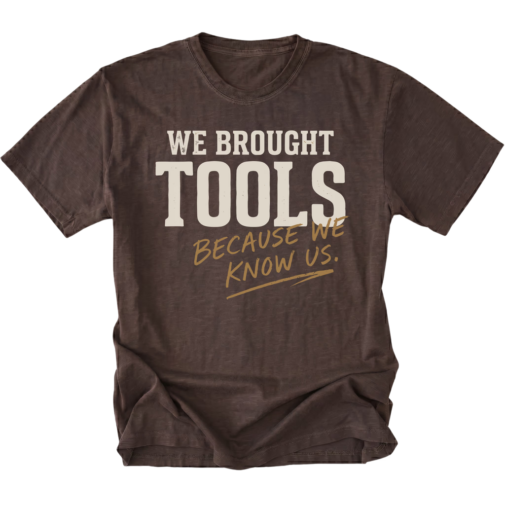A dark brown heather t-shirt with a distressed graphic that reads 'WE BROUGHT TOOLS BECAUSE WE KNOW US.' in cream and tan lettering, displayed on a solid white background.