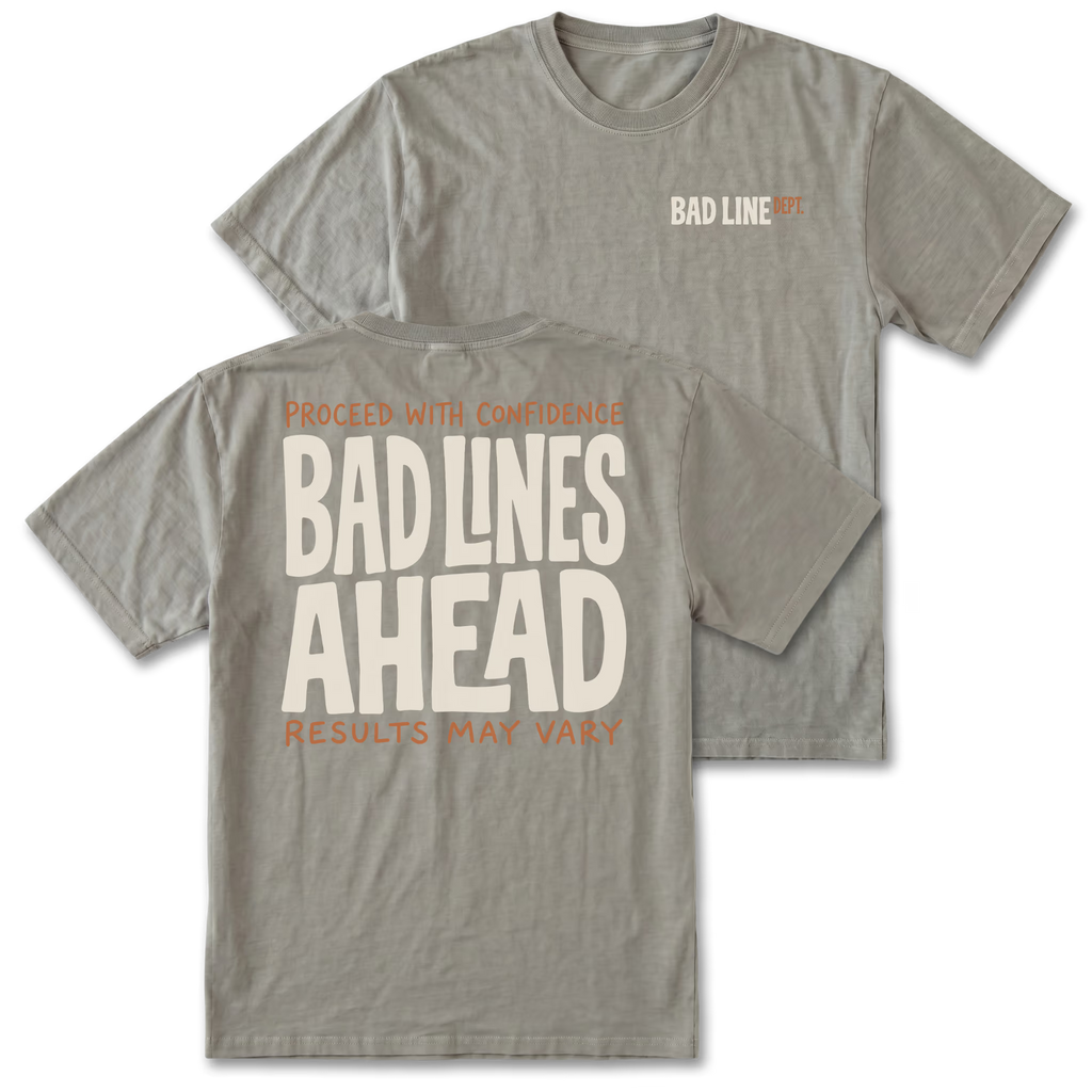 Two gray graphic t-shirts shown on a white background, with the front and back views overlapping. The front features a small 'BAD LINE ART' logo on the left chest, and the back features a large graphic print reading 'PROCEED WITH CONFIDENCE BAD LINES AHEAD RESULTS MAY VARY' in cream and orange lettering.