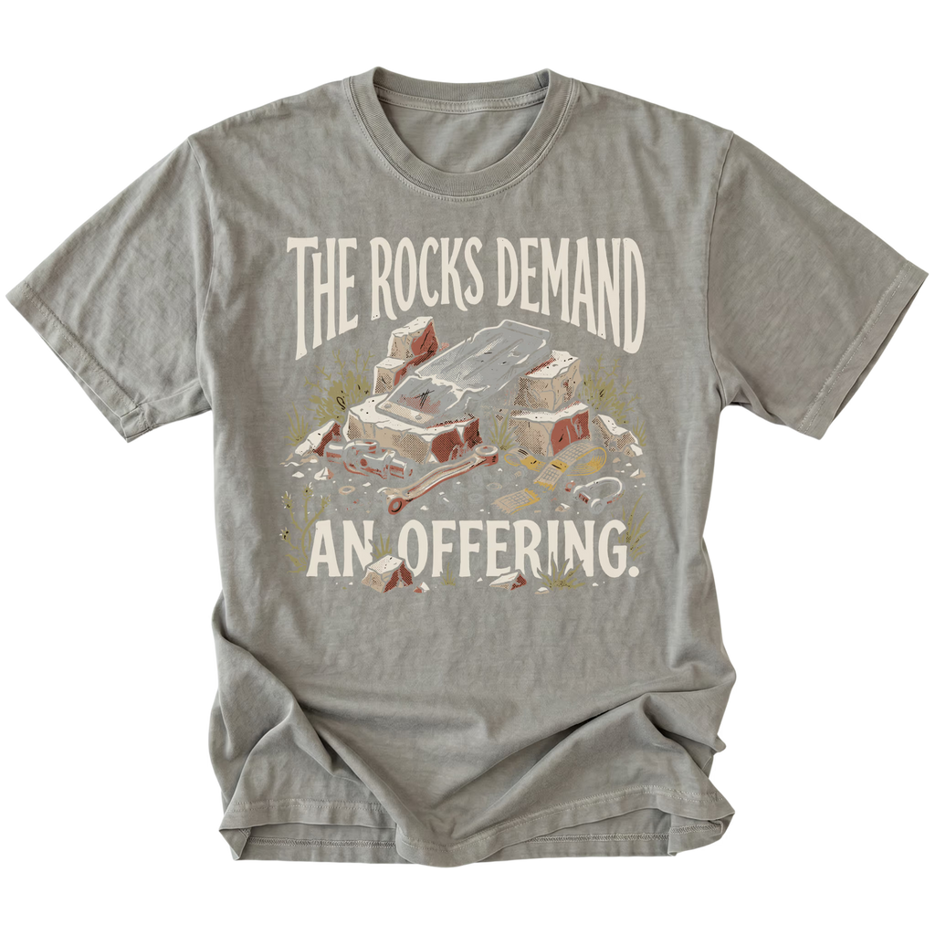 A gray crewneck t-shirt featuring a humorous graphic print of a pile of rocks and a bone with the text 'THE ROCKS DEMAND AN OFFERING.' in a distressed cream font.