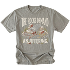 A gray crewneck t-shirt featuring a humorous graphic print of a pile of rocks and a bone with the text 'THE ROCKS DEMAND AN OFFERING.' in a distressed cream font.