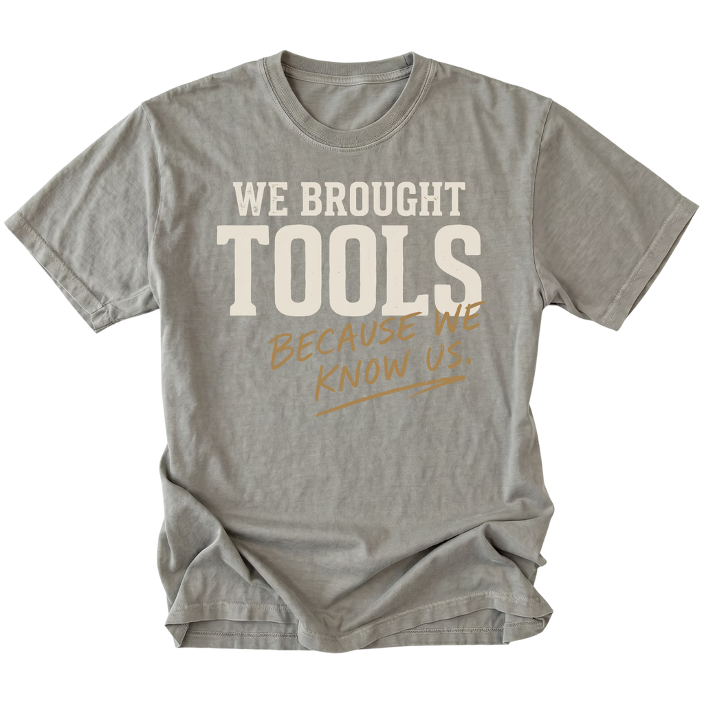 A gray short-sleeve t-shirt with a graphic text print reading 'WE BROUGHT TOOLS BECAUSE WE KNOW US.' in cream and tan lettering, displayed flat against a solid white background.