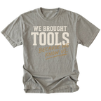 A gray short-sleeve t-shirt with a graphic text print reading 'WE BROUGHT TOOLS BECAUSE WE KNOW US.' in cream and tan lettering, displayed flat against a solid white background.