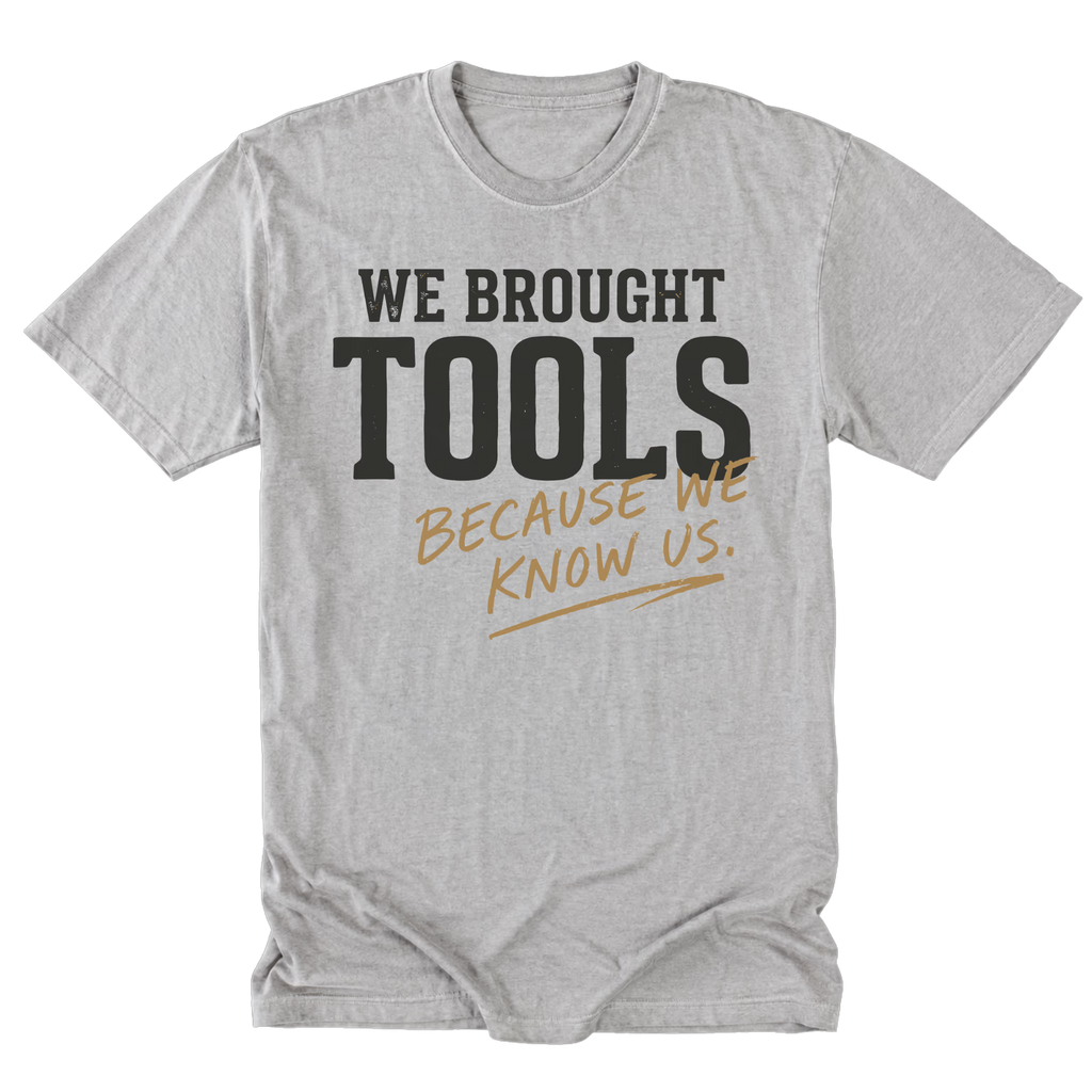 A light steel gray crewneck t-shirt with a graphic print that reads 'WE BROUGHT TOOLS BECAUSE WE KNOW US.' in black and tan lettering, displayed flat against a white background.