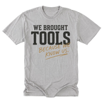 A light steel gray crewneck t-shirt with a graphic print that reads 'WE BROUGHT TOOLS BECAUSE WE KNOW US.' in black and tan lettering, displayed flat against a white background.