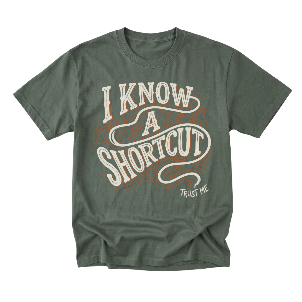 An military green short-sleeve t-shirt with a vintage-style graphic reading 'I KNOW A SHORTCUT' in cream and brown, with 'TRUST ME' printed below in white.