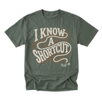 An military green short-sleeve t-shirt with a vintage-style graphic reading 'I KNOW A SHORTCUT' in cream and brown, with 'TRUST ME' printed below in white.