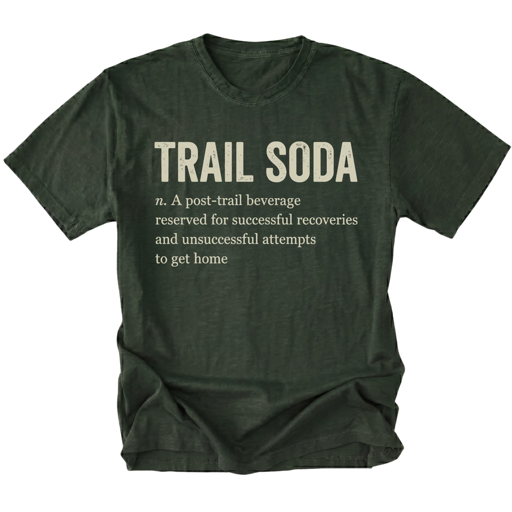 A moss green t-shirt with a humorous definition of 'Trail Soda' printed in a distressed cream-colored font on the front, displayed against a solid white background.