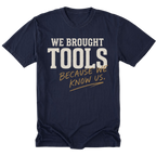 A navy blue short-sleeve t-shirt with a graphic text print reading 'WE BROUGHT TOOLS BECAUSE WE KNOW US.' in white and gold lettering, displayed on a clean white background.