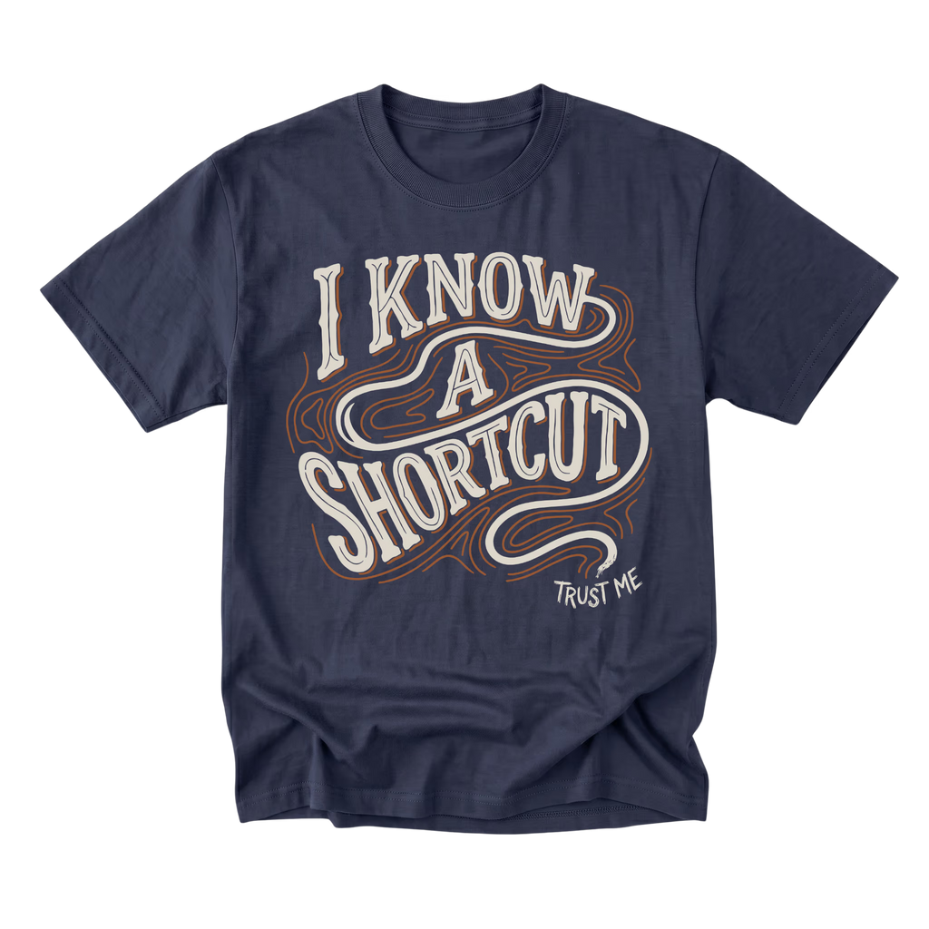 A navy blue short-sleeve t-shirt with a retro-style graphic on the front reading 'I KNOW A SHORTCUT' in white and orange wavy lettering, with 'TRUST ME' printed below in white.