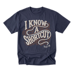 A navy blue short-sleeve t-shirt with a retro-style graphic on the front reading 'I KNOW A SHORTCUT' in white and orange wavy lettering, with 'TRUST ME' printed below in white.