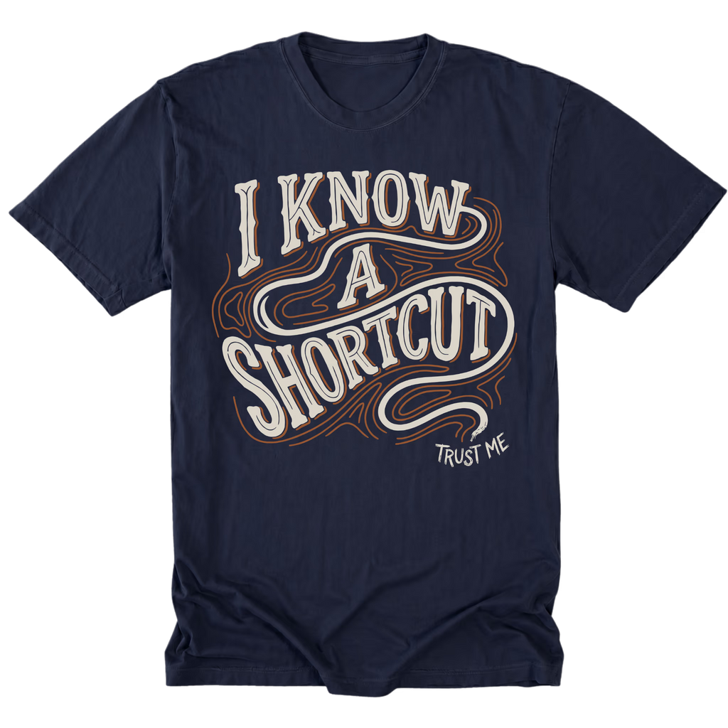 A navy blue short-sleeve t-shirt with a graphic on the front that reads 'I KNOW A SHORTCUT' in white and orange wavy lettering, with 'TRUST ME' written below in white.