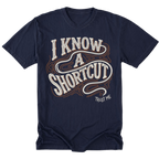 A navy blue short-sleeve t-shirt with a graphic on the front that reads 'I KNOW A SHORTCUT' in white and orange wavy lettering, with 'TRUST ME' written below in white.