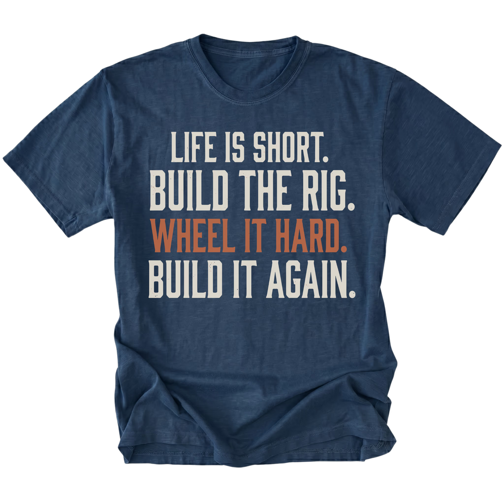 A navy blue short-sleeve t-shirt with a distressed white and orange text graphic on the front reading 'LIFE IS SHORT. BUILD THE RIG. WHEEL IT HARD. BUILD IT AGAIN.'