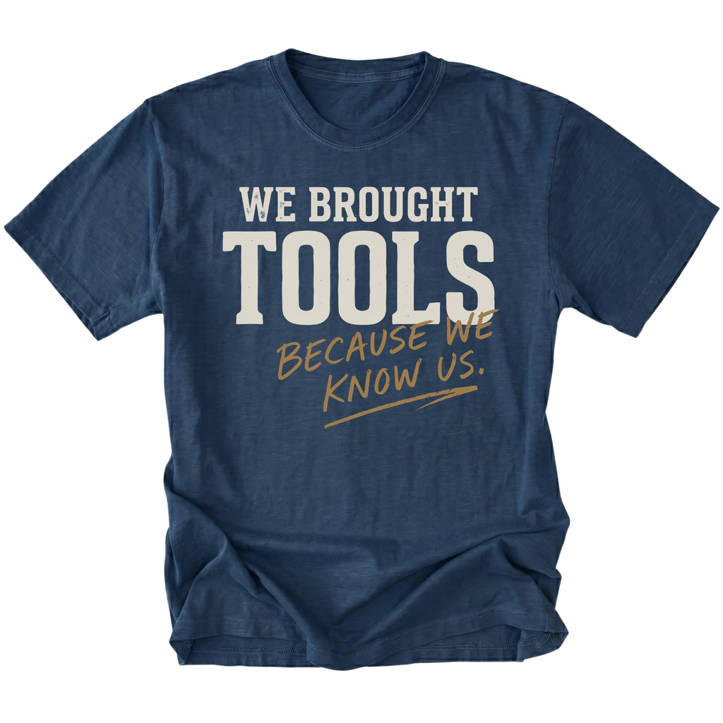 A navy blue short-sleeve t-shirt with a graphic text print reading 'WE BROUGHT TOOLS BECAUSE WE KNOW US.' in white and gold lettering, displayed on a clean white background.