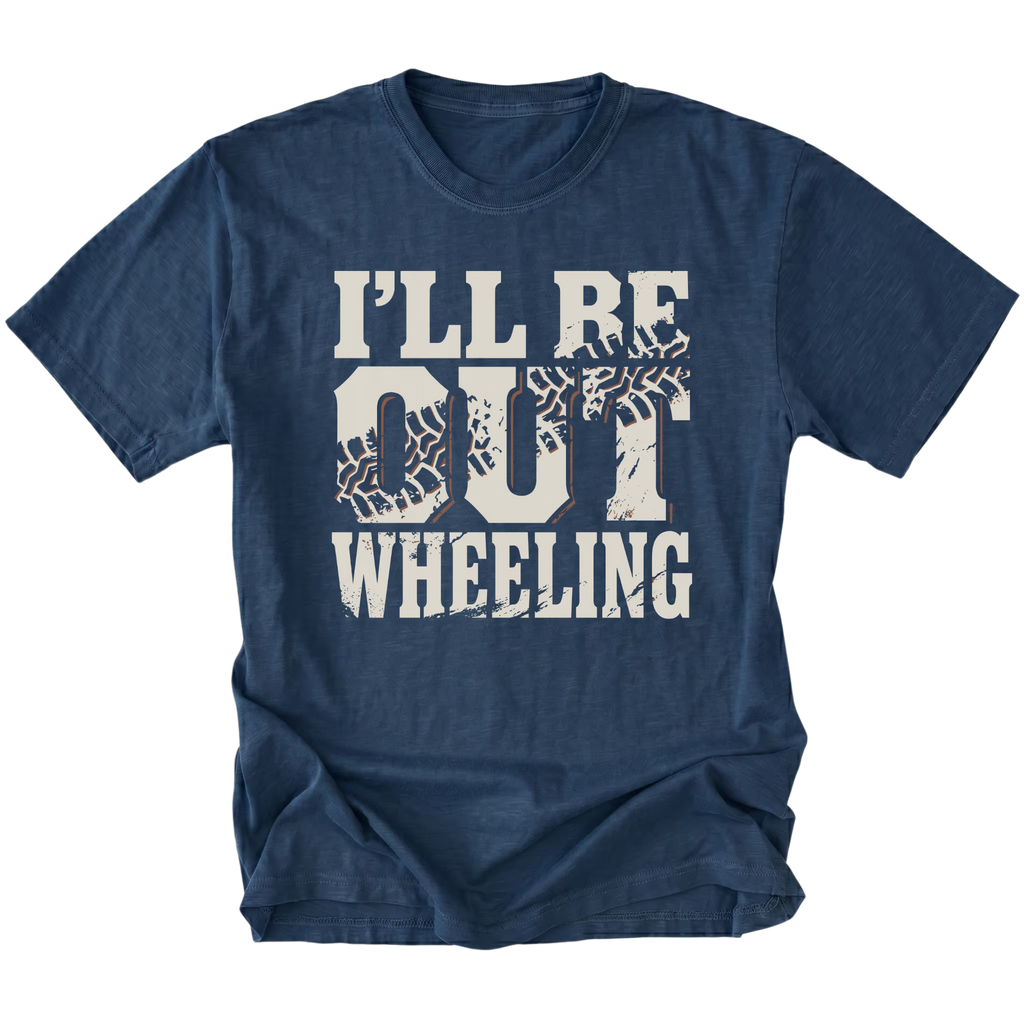 A navy blue short-sleeve t-shirt with a distressed white graphic print that reads 'I'LL BE OUT WHEELING' with tire tracks integrated into the letters, shown on a plain white background.