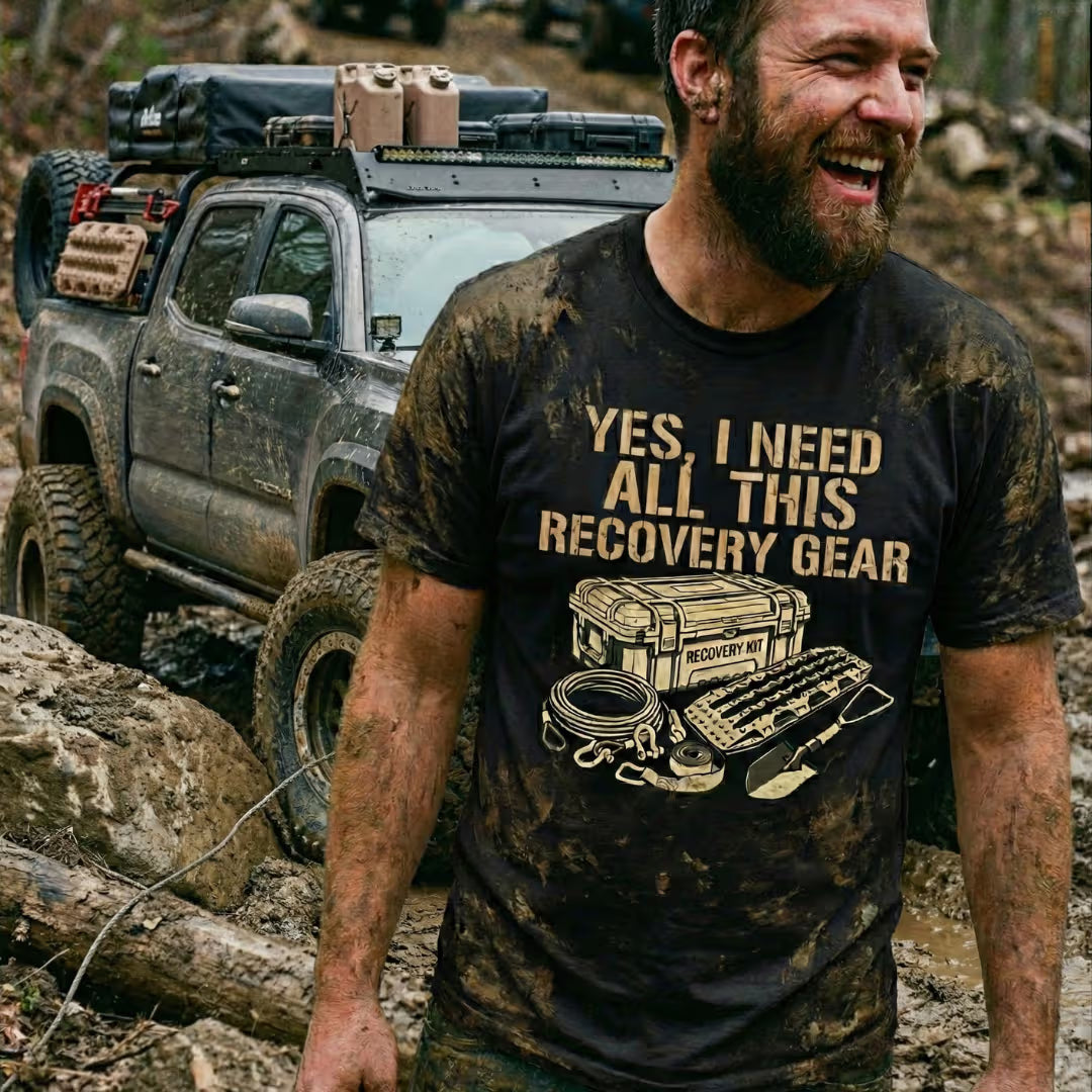 Mud-covered off-road driver wearing a black Trail Snark t-shirt reading ‘Yes, I Need All This Recovery Gear’ with recovery kit graphic, standing in front of a lifted Toyota Tacoma on a rocky trail.